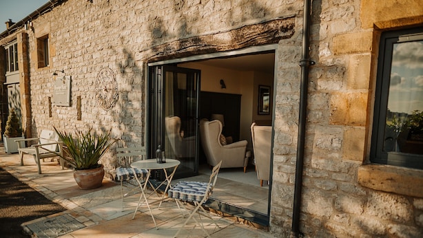 A stone building with a patio area featuring a table, chairs, and a potted plant. - Luxury Rural Holiday Cottage With Log Burner (Buxton)