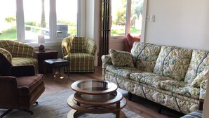 A cozy living room with a sofa, armchairs, and a coffee table. Large windows offer a view of the outdoors. - Bright and Cheerful Lake Front Beach Cottage (Cedar Grove)