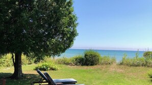 A wooden deck with a patterned rug, a bench, and two lounge chairs facing a grassy area with a view of the sea. - Bright and Cheerful Lake Front Beach Cottage (Cedar Grove)