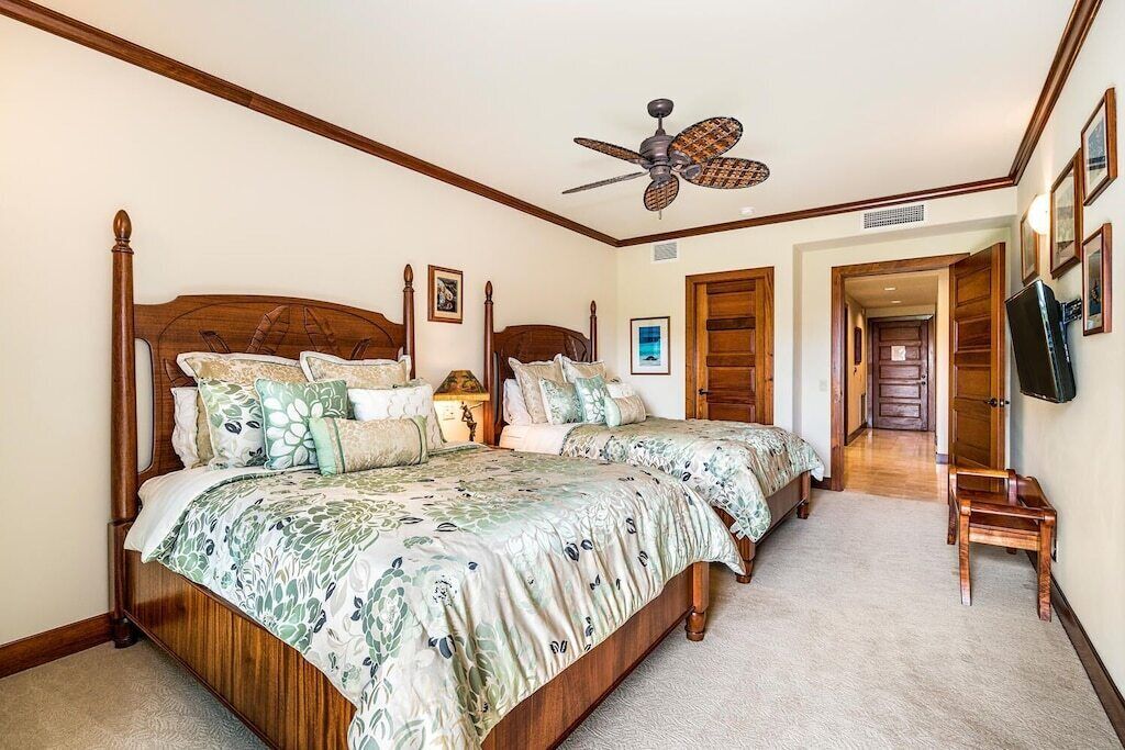 Quarto com duas camas, cabeceiras de madeira, ventoinha no teto e televisão na parede.