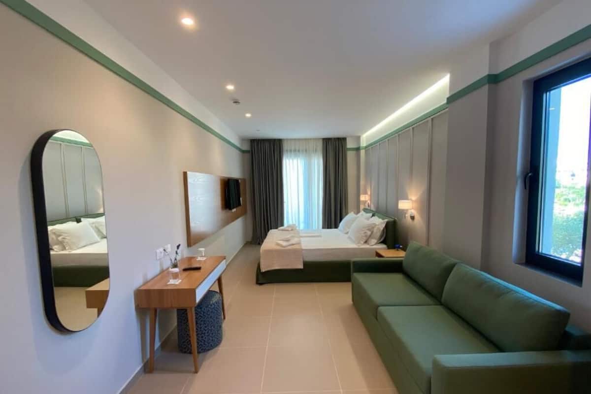 A modern hotel room with a bed, a green sofa, a small table with a lamp, and a large window.