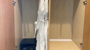 A walk-in closet with wooden shelving, a robe hanging on a rack, a black suitcase, and a pair of shoes on the floor. - Olive Hotel Ksamil (Ksamil)