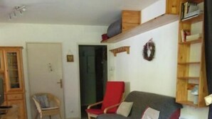 Uma sala de estar acolhedora com um sofá de madeira, uma poltrona vermelha e uma mesa de centro de madeira. Há uma estante com livros e uma grinalda decorativa na parede.