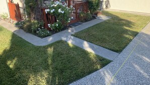 Una casa residencial con jardín delantero, terraza de madera y un pequeño jardín con flores blancas.