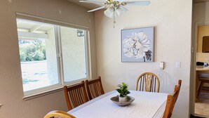 Comedor con una mesa dispuesta para seis, ventilador de techo, ventana con vistas y un cuadro de flores colgado en la pared.