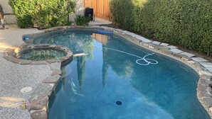 Una piscina con borde de piedra, rodeada por un jardín con diversas plantas y una valla de madera al fondo.