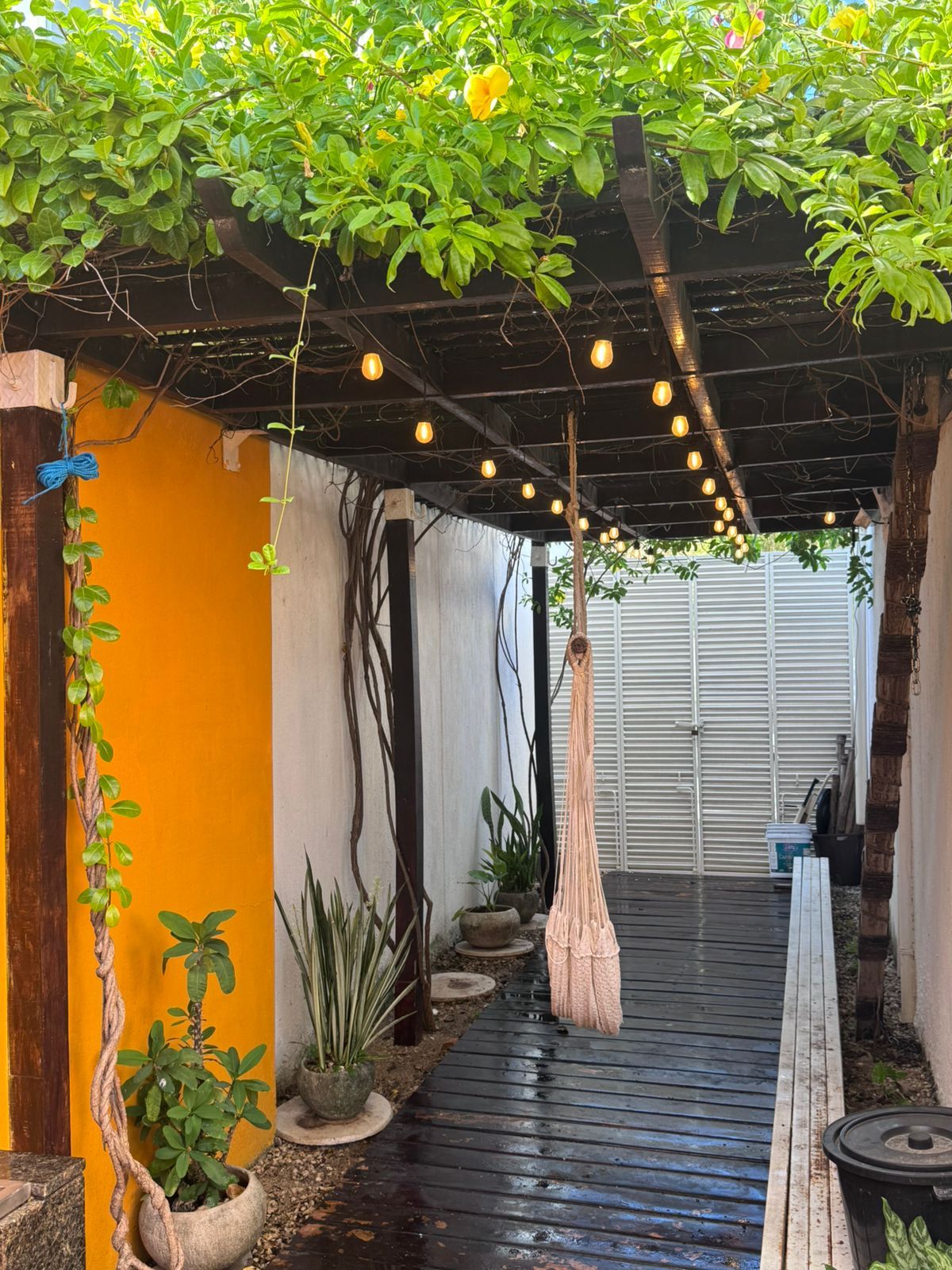 A covered outdoor area with a wooden floor, potted plants, and hanging lights.
