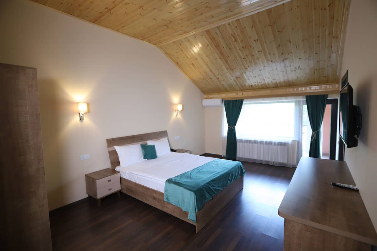 A bedroom with a wooden ceiling, a bed with a green bedspread, a nightstand, a bedside table, and a TV mounted on the wall.