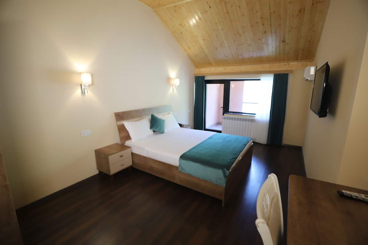 A bedroom with a wooden ceiling, a bed with a teal blanket, a nightstand, a TV mounted on the wall, and a window with curtains.