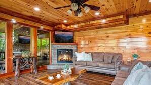 A cozy cabin interior with a stone fireplace, wooden walls, a leather sofa, and a dining table with a rustic centerpiece. - Large Luxury Cabin Near Pigeon Forge | Indoor Pool, Theater, Sleeps 56 (Sevierville)