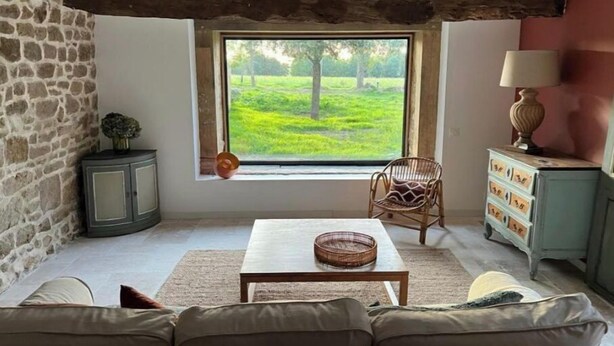 Ein gemütliches Zimmer mit Steinwand, Holzboden, einem großen Fenster mit Blick auf Grünflächen, einem Korbwägelchen auf einem Tisch und einer Lampe auf einem Schrank.