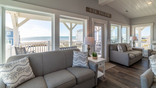 A spacious living area with a view of the beach, a sign reading "OAK ISLAND," and a balcony with outdoor furniture.