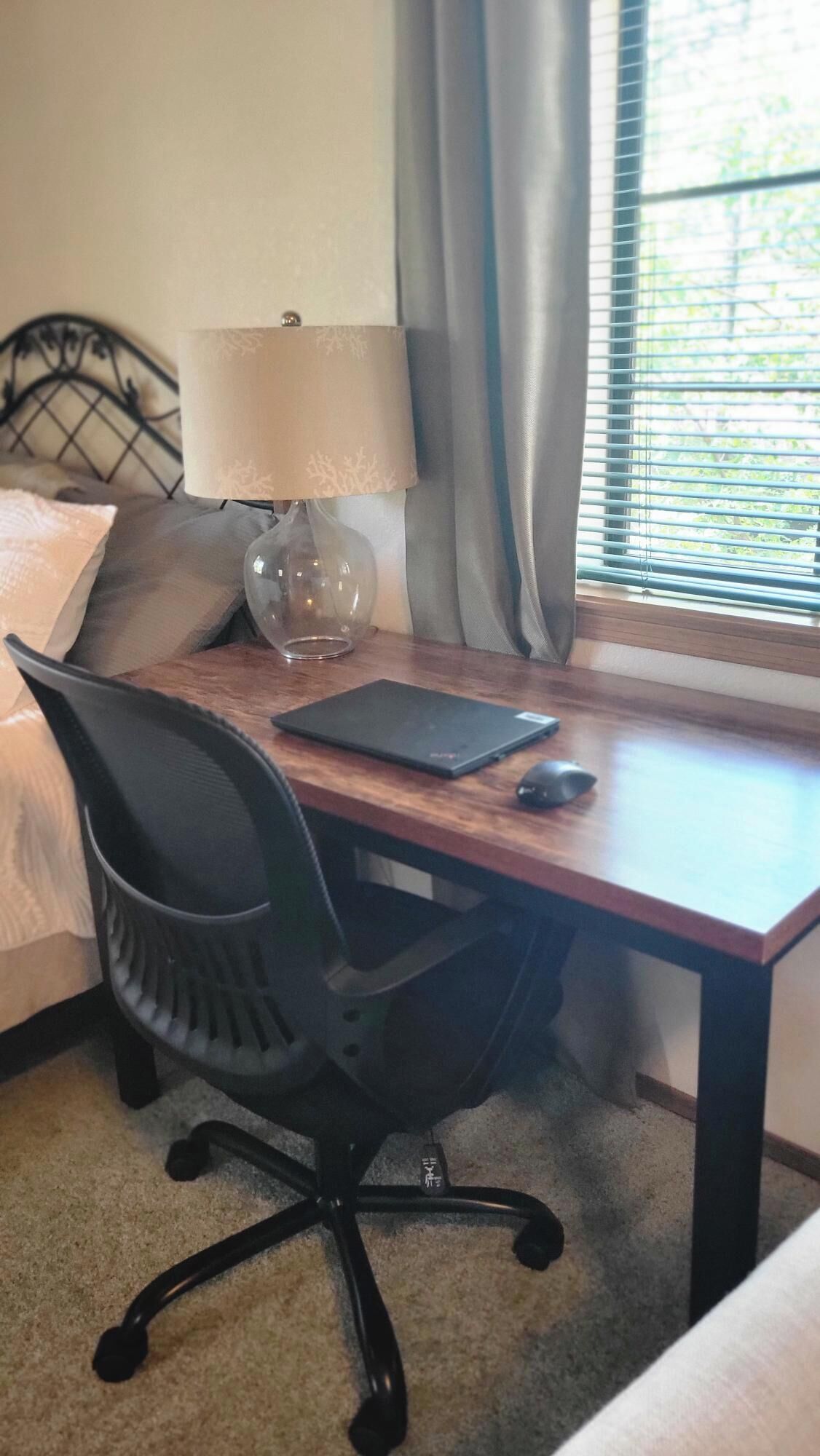 Quarto com cama, escrivaninha com laptop, cadeira, luminária e janela com cortinas.