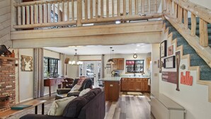 A spacious living area with a dark brown sofa, a wooden bench, and a brick fireplace. A kitchen with wooden cabinets and a large island is visible in the background. - Tranquil Pinetop Getaway! w/Firepit, Grill, Deck, Pet-Friendly, Near Ski & Golf! (Pinetop)