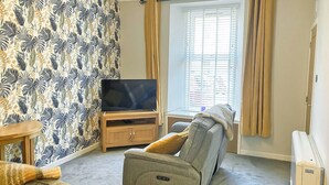 A living room with a sofa, a television on a wooden stand, a window with blinds, and a patterned wallpaper. - A lovely hideaway in the heart of historic Crieff. (Crieff)