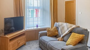 A living room with a television on a wooden stand, a grey sofa with cushions, a window with blinds, and a door. - A lovely hideaway in the heart of historic Crieff. (Crieff)