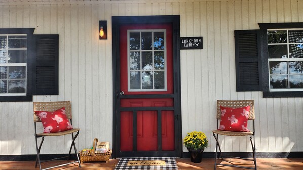 En entré till stugan med en röd dörr, två flätade fåtöljer med röda kuddar, en välkomstmatta och en skylt med texten ”Longhorn Cabin”.