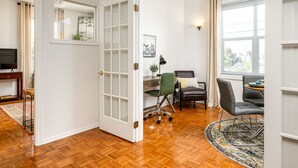 Ein Zimmer mit Holzboden, Schreibtisch mit Stuhl, Esstisch mit Stühlen, einem Fernseher und einem Fenster mit Vorhängen.