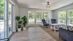 A bright sunroom with large windows, a wooden floor, a table with books, two chairs, and a sofa. - Fun Getaway Lakehouse in the Hudson Valley (Craryville)