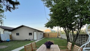 A patio with a wooden table and chairs, a small garden, and a house in the background. - Main Central Avenue Basement Studio (Saskatoon)