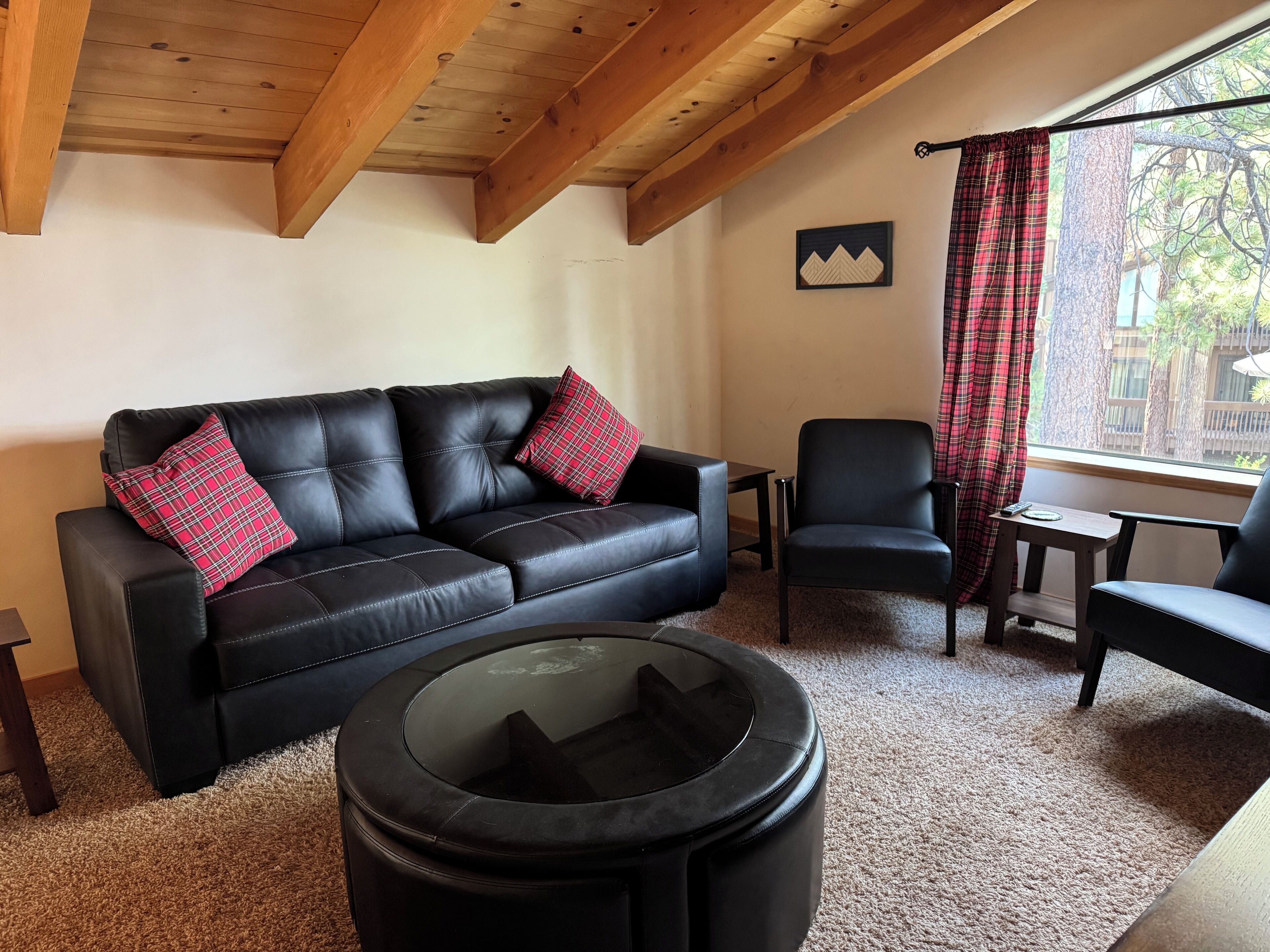 Una sala de estar con un sofá de cuero negro, dos almohadones a cuadros rojos, un sillón negro, una pequeña mesa de madera y una mesa de centro de vidrio.