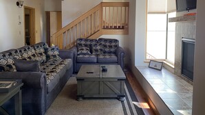 A spacious living room with a large gray sectional sofa, a wooden coffee table, and a fireplace. A wooden staircase with a railing is visible in the background.