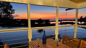 Un espace extérieur abrité, avec une table et des chaises en bois, offrant une vue sur un plan d’eau au coucher du soleil.