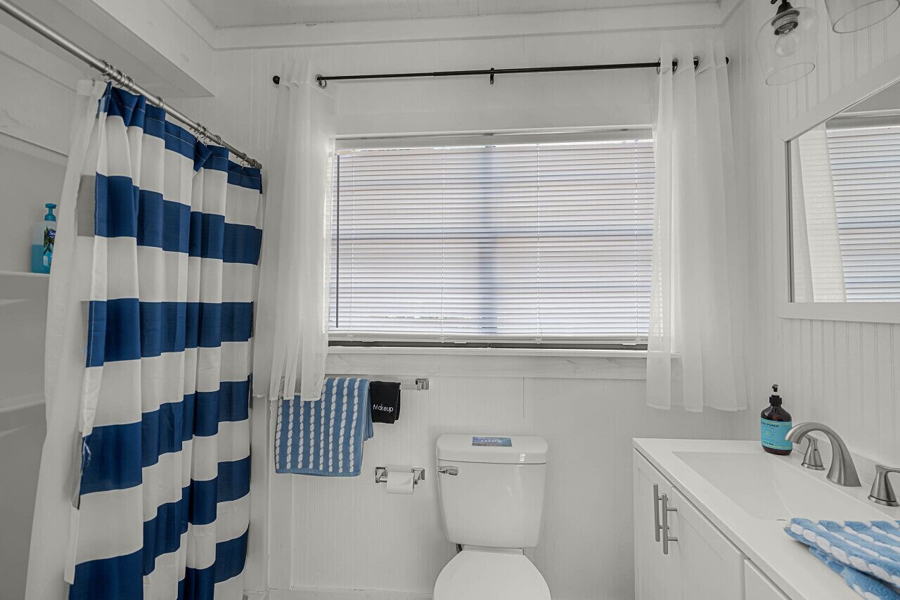 Une salle de bain avec des toilettes blanches, un lavabo avec un miroir et une fenêtre avec des stores.