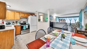 Cocina compacta con armarios de madera, un refrigerador y un microondas. Hay una zona de comedor con mantel a cuadros y sillas.