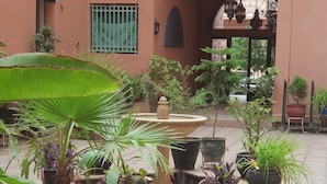Een binnentuin met plantenpotten, een fontein en een gebouw met een groen raam en een rondboogvormige ingang.