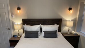 A neatly made bed with white linens and two dark gray pillows, flanked by two bedside tables with lamps, one of which has a digital clock displaying 4:19.