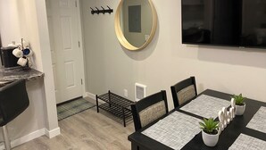 A dining area with a black table, four chairs, a round mirror, and a flat-screen TV on the wall.