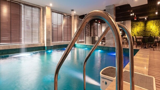 An indoor swimming pool with a metal ladder, wooden deck, and a seating area with tables and chairs. - Hotel Snowland Srinagar (Srinagar)