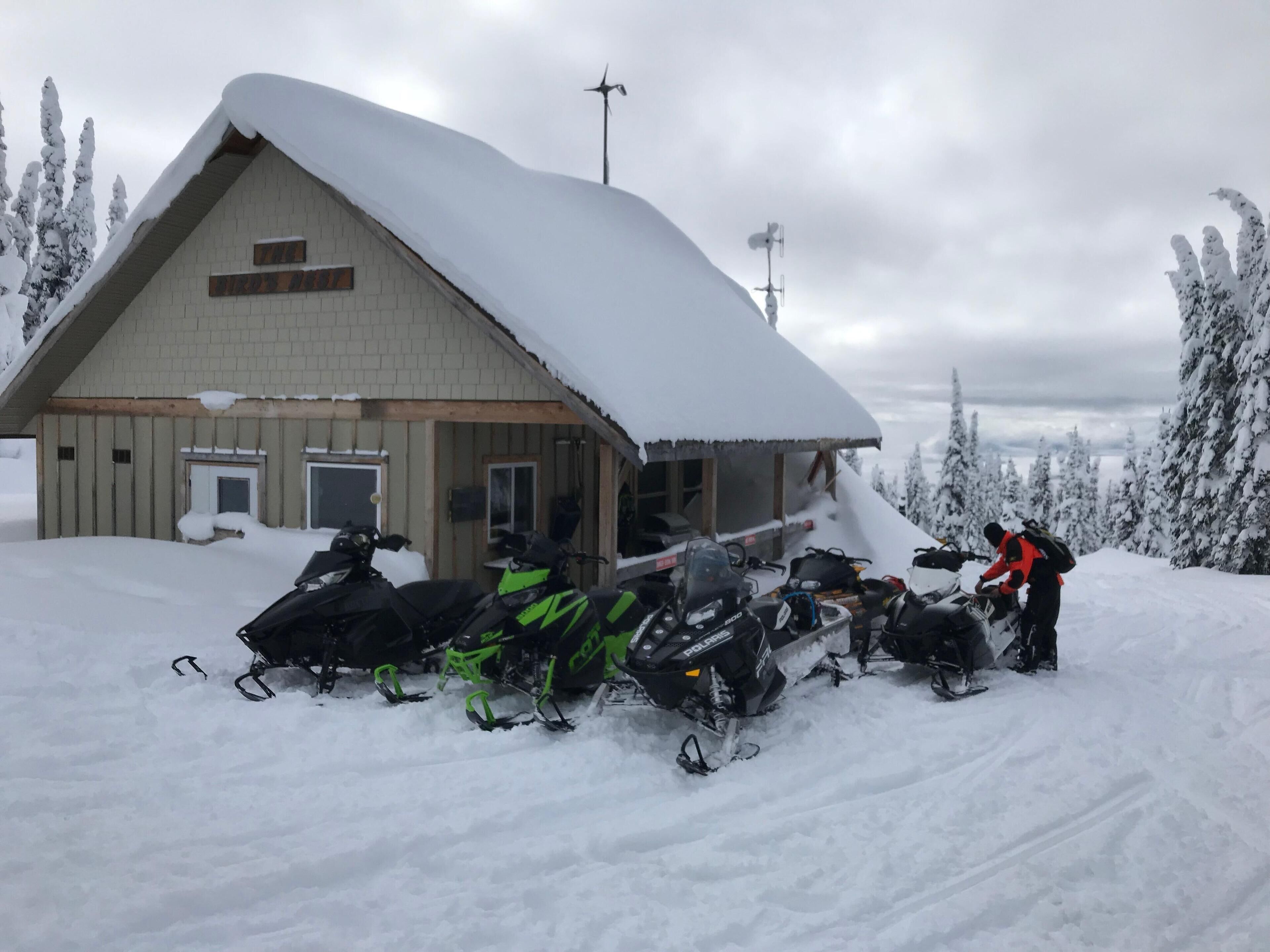 Un chalet enneigé, avec une façade en bois et un toit à forte pente. Plusieurs motoneiges sont garées devant.