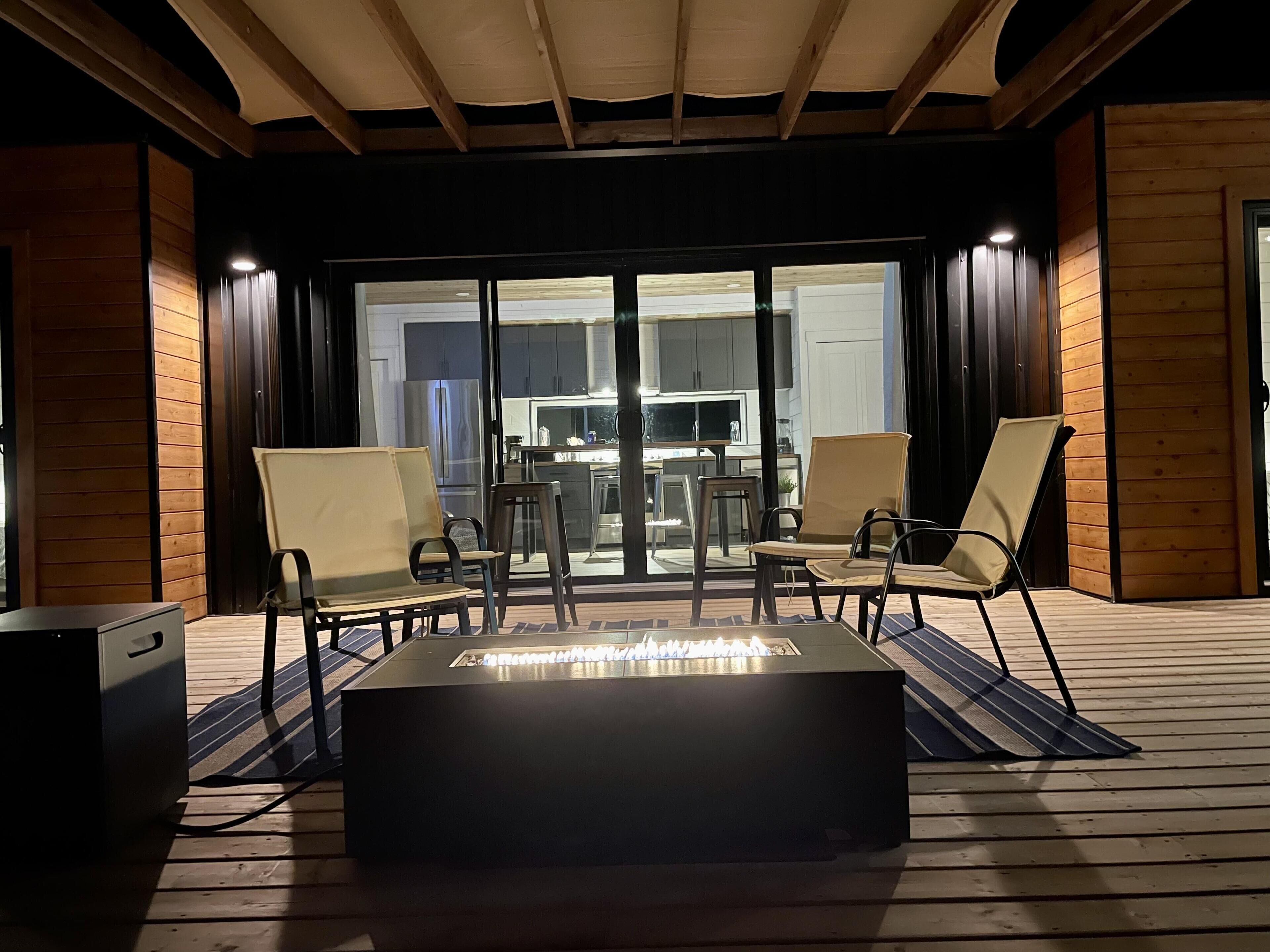 Une terrasse extérieure dotée d’un foyer, de deux chaises longues et offrant une vue sur la cuisine à travers des portes vitrées.