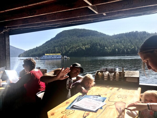 L’intérieur d’un bateau, avec une table dressée pour le repas, une vue sur une île boisée et un ferry en arrière-plan.