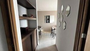 Un couloir étroit, avec un coin cuisine sur la gauche, un espace repas avec des chaises et une table au centre, et un mur agrémenté d’éléments décoratifs sur la droite.