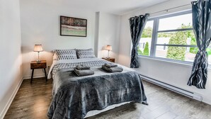 Ein Schlafzimmer mit einem Bett, zwei Nachttischen, einem Fenster mit Vorhängen, einem Bild an der Wand und Blick ins Grüne.
