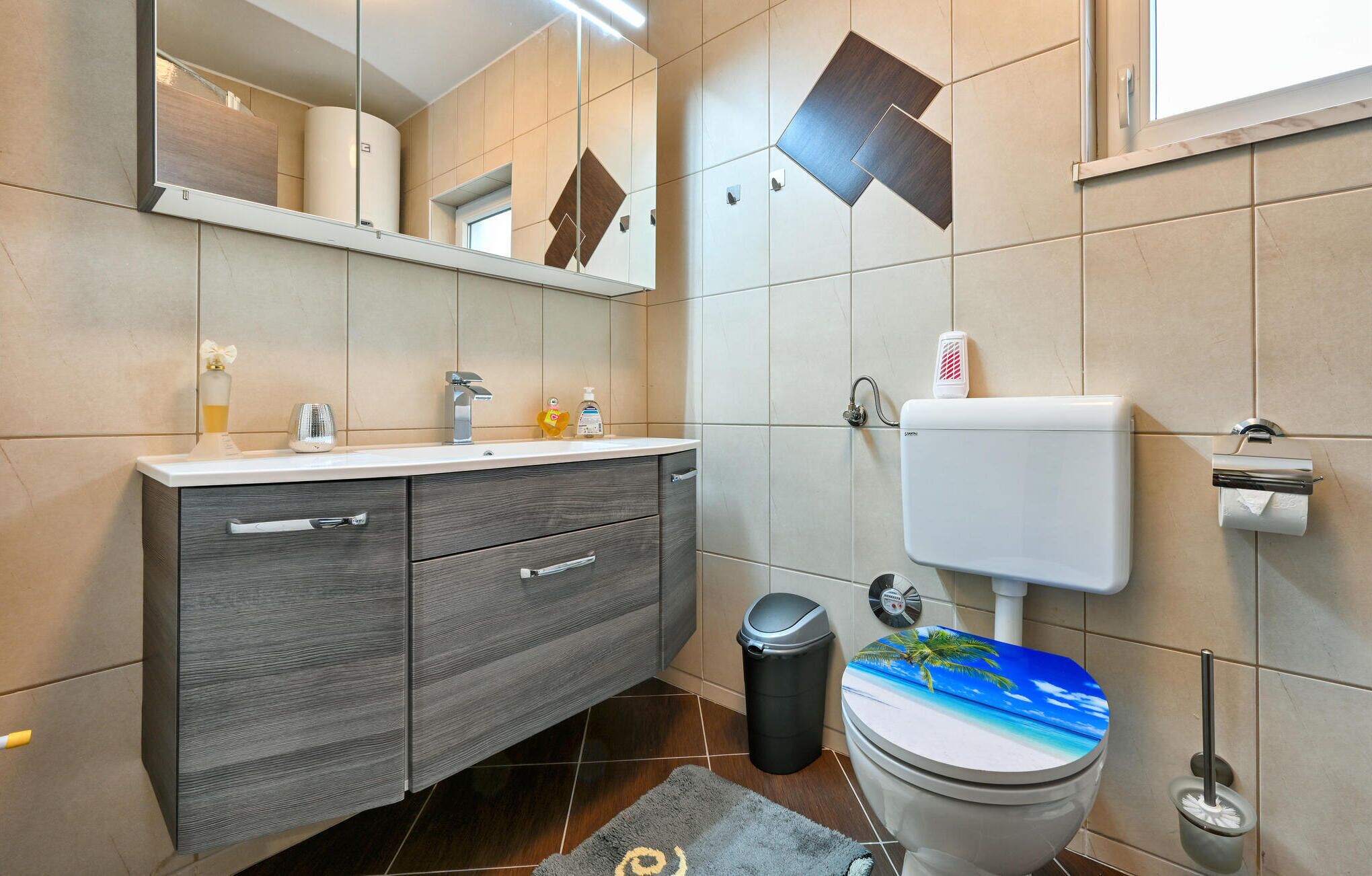 Un baño moderno con un mueble de tocador gris, un inodoro con tapa decorada con motivos playeros y un cubo de basura.