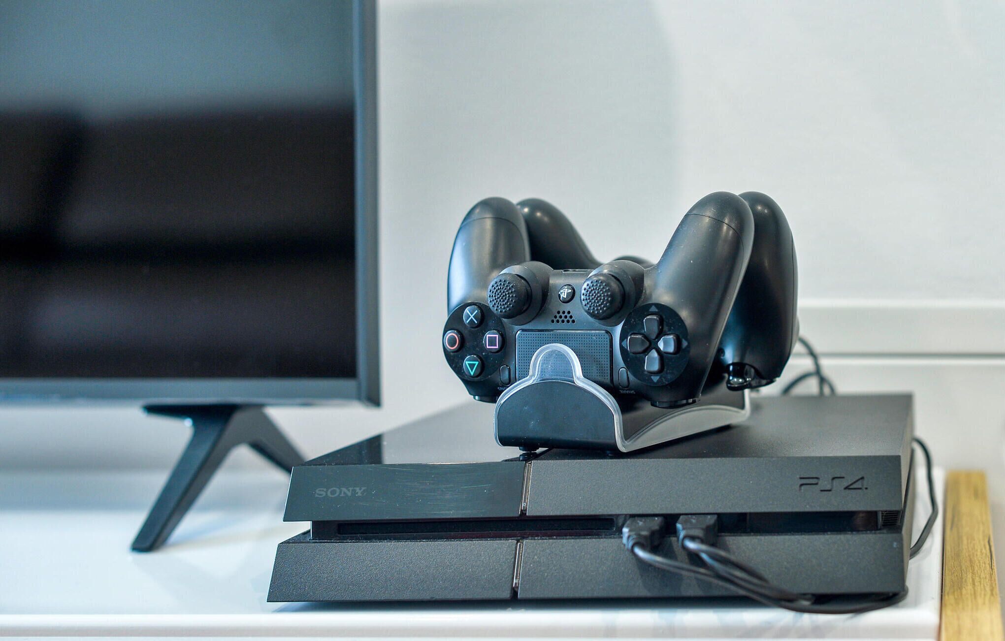 Een PlayStation 4 console met een zwarte controller erop, geplaatst op een wit oppervlak met een monitor op de achtergrond.