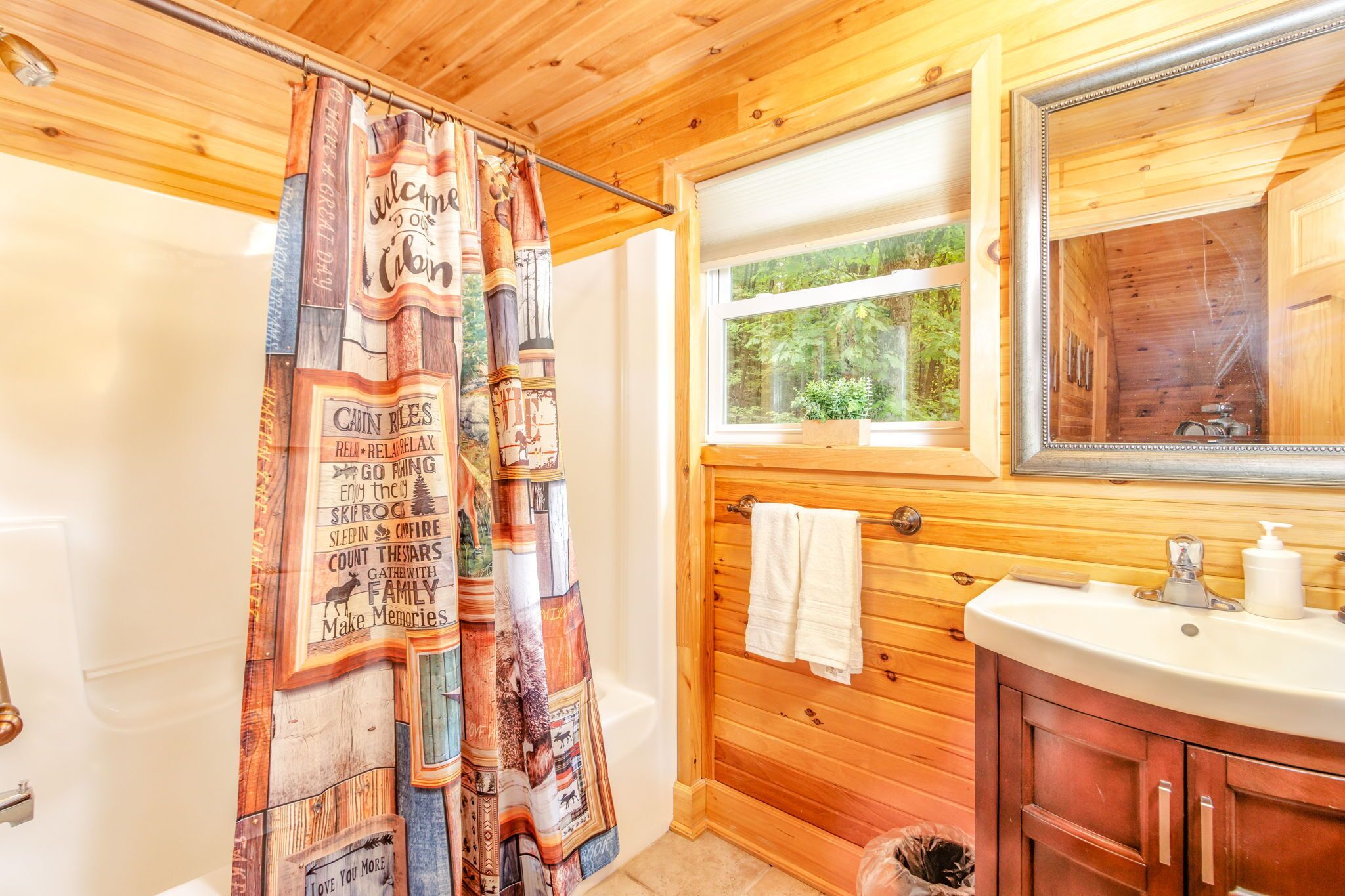 A cabin-style bathroom with a wooden interior, a shower curtain with text, a window with a view of greenery, and a sink with a mirror.