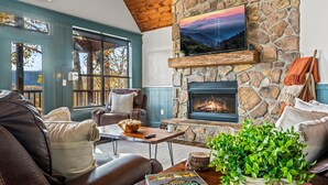 Un salón acogedor con chimenea de piedra, mobiliario de cuero y un amplio ventanal con vistas a un bosque.