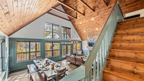 Un comedor amplio con techo alto y vigas de madera, una escalera con barandilla azul claro y ventanas grandes que ofrecen vistas al exterior.