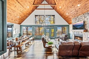 A spacious loft with a high ceiling, wooden beams, a stone fireplace, and large windows offering views of the outdoors. - Indoor Pool 💦 Theater 🎬 Game Room 🎮 | Sleeps 17 | 1 Mile to Dollywood (Sevierville)