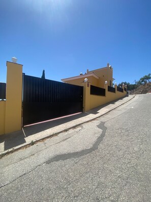 Un edificio amarillo con una reja negra, cielo despejado y un árbol al fondo.