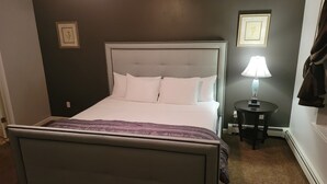 Een net opgemaakt bed met witte lakens en een paarse plaid, een nachtkastje met een lamp, en twee ingelijste foto's aan de muur.