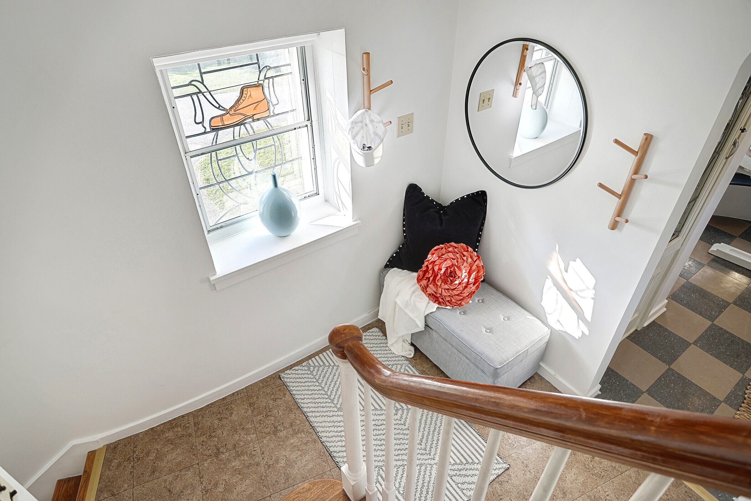 A staircase with a wooden handrail, a white wall, a window with a view of a garden, a round mirror, and a grey bench with a red flower.