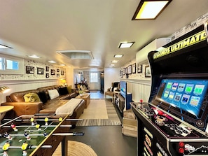 A cozy living room with a foosball table, a comfortable sofa, and a television displaying various game options. - Iconic Haines Shoe House - Hot Tub, Firepit, Foosball & 300 Game Arcade Machine! (Yorkana)