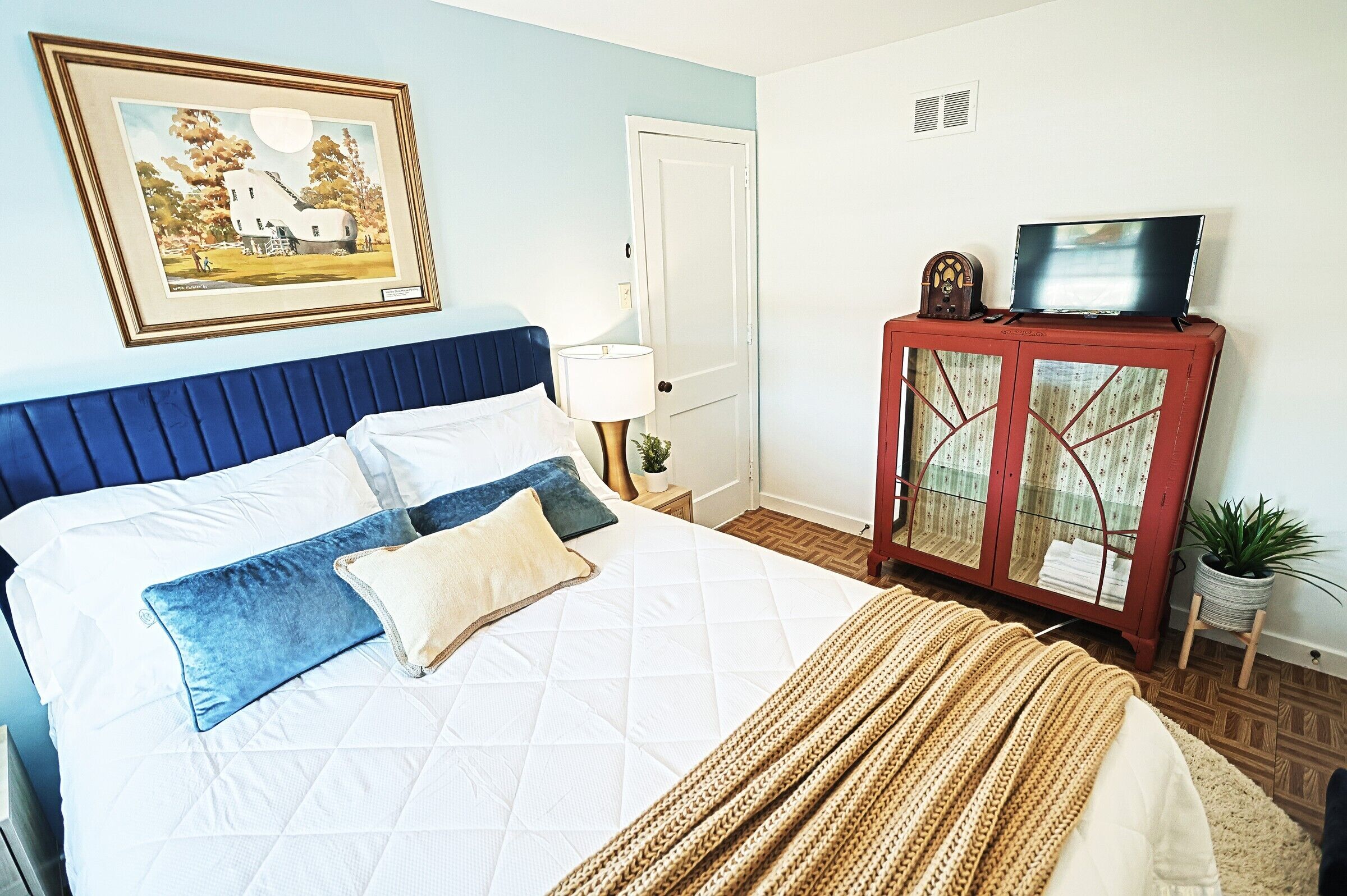 A neatly made bed with a blue headboard, a framed picture on the wall, a television on a cabinet, and a small plant on a side table.