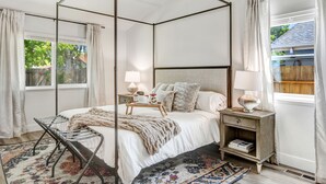 Uma cama de dossel com estrutura de madeira, um criado-mudo, uma mesinha com abajur e um tapete estampado.